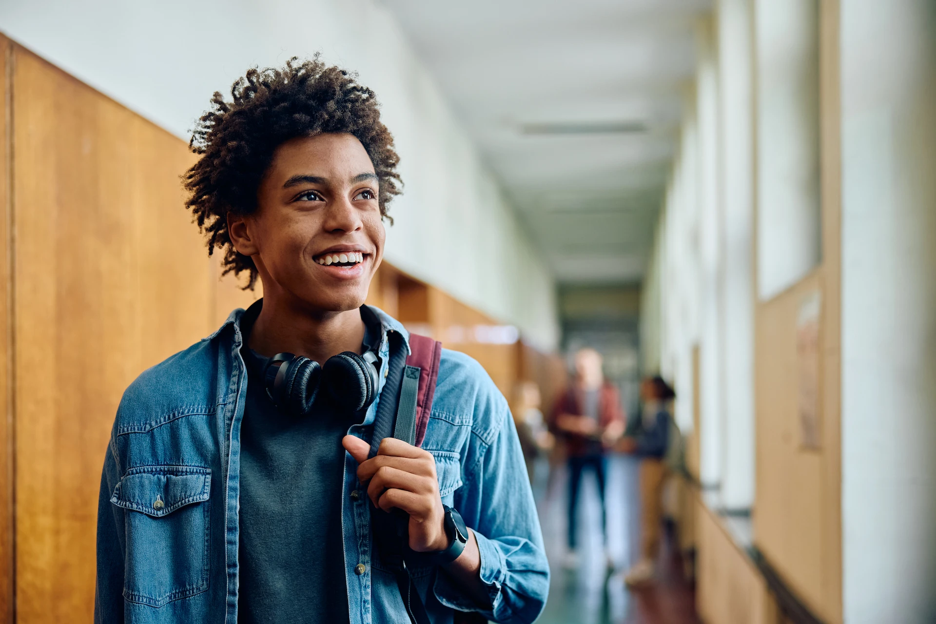 A smiling teenager with curly hair, wearing headphones around their neck and a backpack, stands in a school hallway, ready to seek business tutoring help among blurred students and windows in the background.
