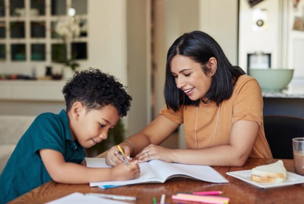 A woman and a young boy sit at a table, smiling and working together on a drawing or homework in a notebook. Colored pencils, paper, and a plate with a sandwich are on the table.