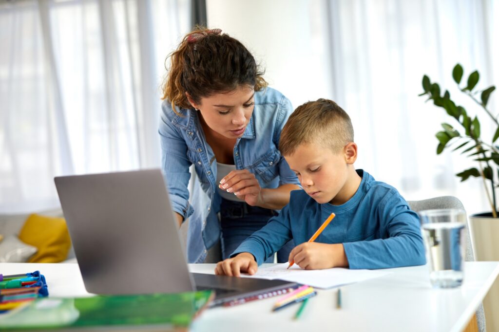 A woman leans over to help a young boy who is writing on paper at a table with a laptop, colored pencils, books, and a glass of water in a bright room with large windows and a plant in the background.