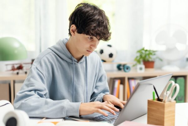 A teenage boy wearing a light blue hoodie sits at a desk typing on a laptop. The background includes books, a plant, a soccer ball, and various stationery items.
