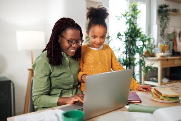 A woman wearing glasses and a green shirt sits at a table using a laptop, smiling, with a young girl in a yellow sweater standing beside her. A plate with a sandwich is on the table. They are in a bright, cozy room.