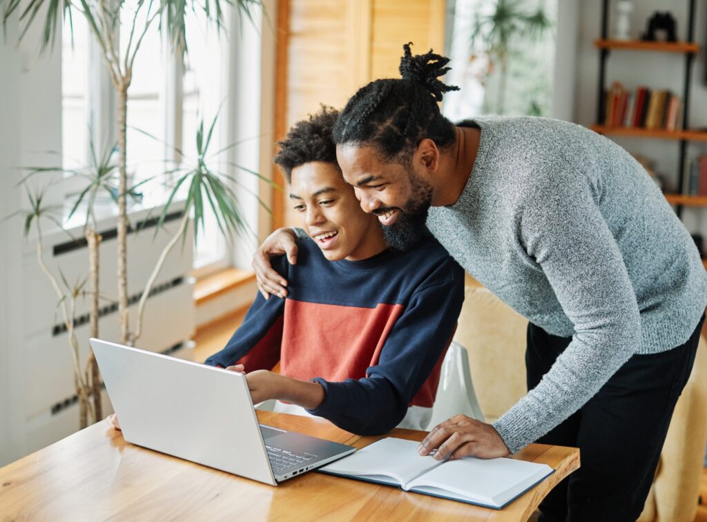 A smiling adult and a teenager sit together at a table, looking at a laptop screen. The adult has one arm around the teens shoulder. An open notebook lies nearby in a bright, cozy room with plants and bookshelves.