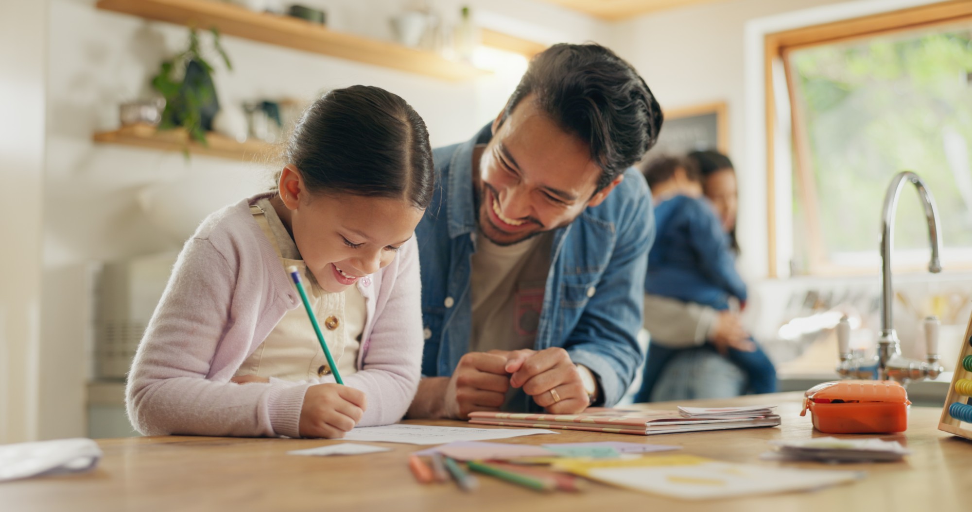 A smiling man helps a young girl with her homework at a kitchen table, while another adult holds a child in the background. The scene is warm and bright, with papers and colored pencils scattered on the table.
