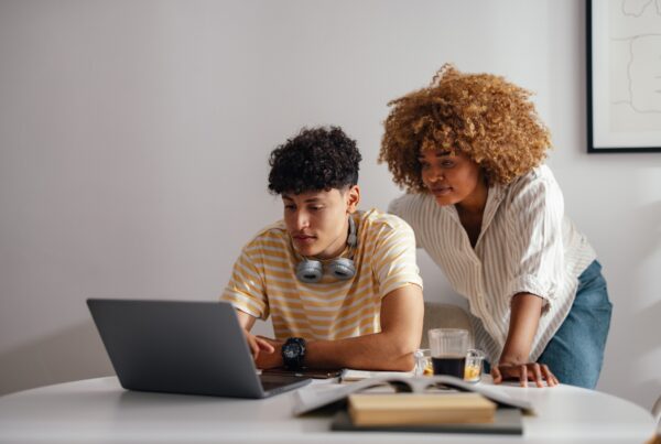 Two young people review something on a laptop at a desk. One sits and types, wearing headphones around his neck; the other stands next to him, leaning over to look at the screen. Books and drinks are on the table.