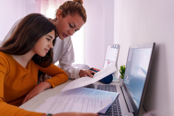 A woman and a teenage girl sit at a desk with a laptop, reading and discussing printed documents together in a bright, cozy room.