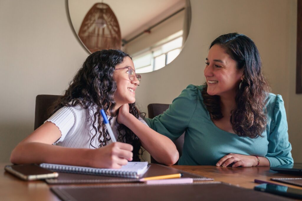A woman and a girl with curly hair sit at a table, smiling at each other. The girl holds a pen over a notebook, while the woman rests her hand on the girls shoulder. They appear to be working or studying together.