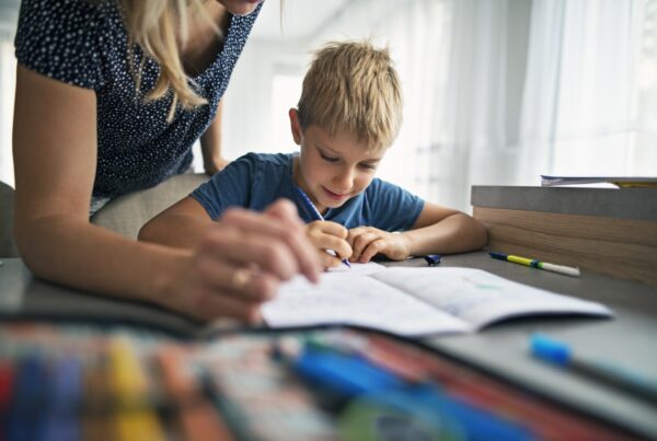A young boy sits at a table, smiling and writing in a notebook, while an adult, partially visible, leans over to help. Colored pencils and markers are spread out on the table in the foreground.